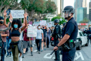 The image shows protesters wearing masks to protect themselves and others from Covid-19. There are cops with helmets and batons on the side of the march. 